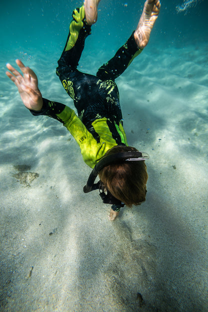 A child in a Waihana Lime Fishbone Steamer wetsuit dives toward a sandy ocean floor in clear turquoise water, wearing a black mask and snorkel.
