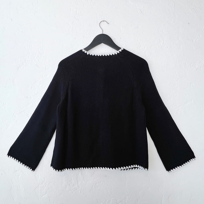 Back side of a black sweater with white trim on a hanger against a white wall.
