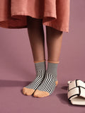 Corbusier Crew socks in Black — graphic op-art black and white stripe with cream rolled cuff and tan heel, worn barefoot under a pink midi skirt with white bow slides nearby