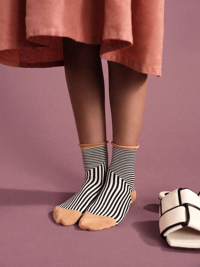 Corbusier Crew socks in Black — graphic op-art black and white stripe with cream rolled cuff and tan heel, worn barefoot under a pink midi skirt with white bow slides nearby