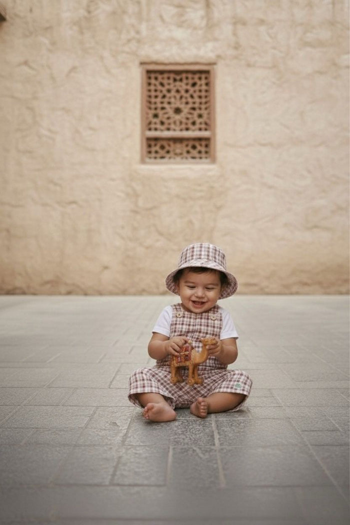 A lifestyle photograph of a smiling baby sitting barefoot and cross-legged on a grey stone-tiled floor. The baby is wearing the Cordoba set, which includes short romper overalls and a matching bucket hat in a red-brown and cream checkered plaid fabric, layered over a simple white short-sleeved t-shirt. The baby is looking down and playing with a small toy camel against a background of a warm, sandy-coloured textured stone wall with an ornate lattice window cutout.