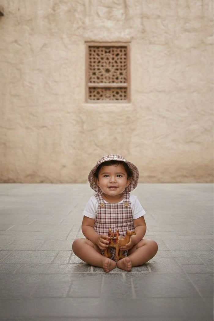 A portrait-style lifestyle photograph of a smiling baby sitting cross-legged and barefoot in the center of a grey stone-tiled floor. The baby is wearing the Cordoba outfit, featuring short romper overalls and a matching bucket hat in a red-brown and cream checkered plaid fabric, worn over a plain white short-sleeved t-shirt. The baby is holding a small toy camel against a backdrop of a warm, sandy-colored textured stone wall with a centrally framed ornate lattice window cutout.