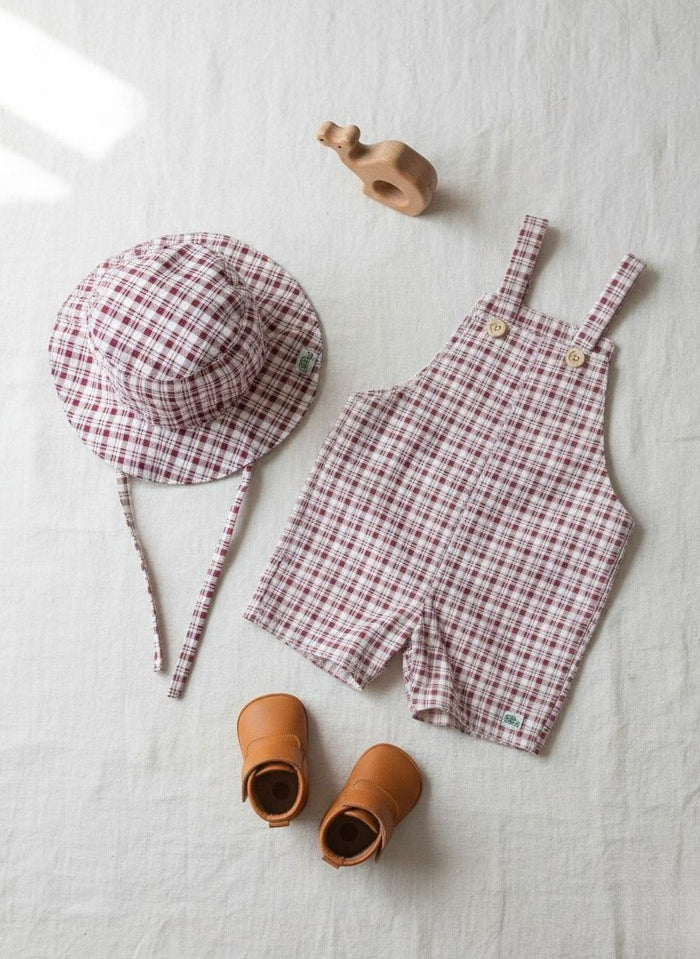 A flat lay photograph of a baby clothing set featuring the Cordoba short romper overalls and a matching bucket hat. Both pieces are made from a red-brown and cream checkered plaid fabric. The overalls feature shoulder straps secured with two small light-colored wooden buttons, while the matching bucket hat includes fabric chin tie straps. The items are styled on a neutral cream linen background next to a pair of tan leather baby booties and a small wooden animal toy.