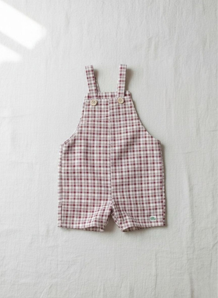 A flat lay photograph of the Cordoba baby short romper overalls, featuring a red-brown and cream checkered plaid pattern. The overalls are detailed with two small, light-colored wooden buttons securing the shoulder straps and a small square green brand label near the right leg hem. It is styled flat and centered on a crisp, neutral white linen background in soft, natural lighting.