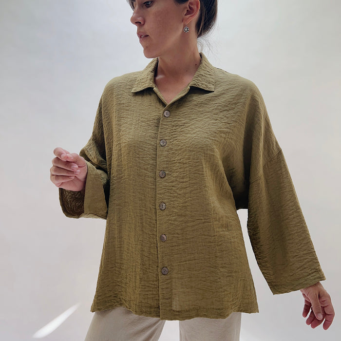 A 3/4 front view of a woman wearing a textured olive green long-sleeve button-up shirt with a relaxed fit, paired with light beige pants, photographed against a neutral light gray background.
