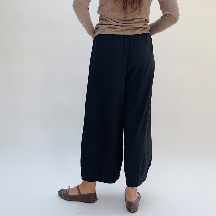A person standing against a white background, wearing black cropped pants with darts at the hemline, a beige top, and dark shoes, viewed from the back