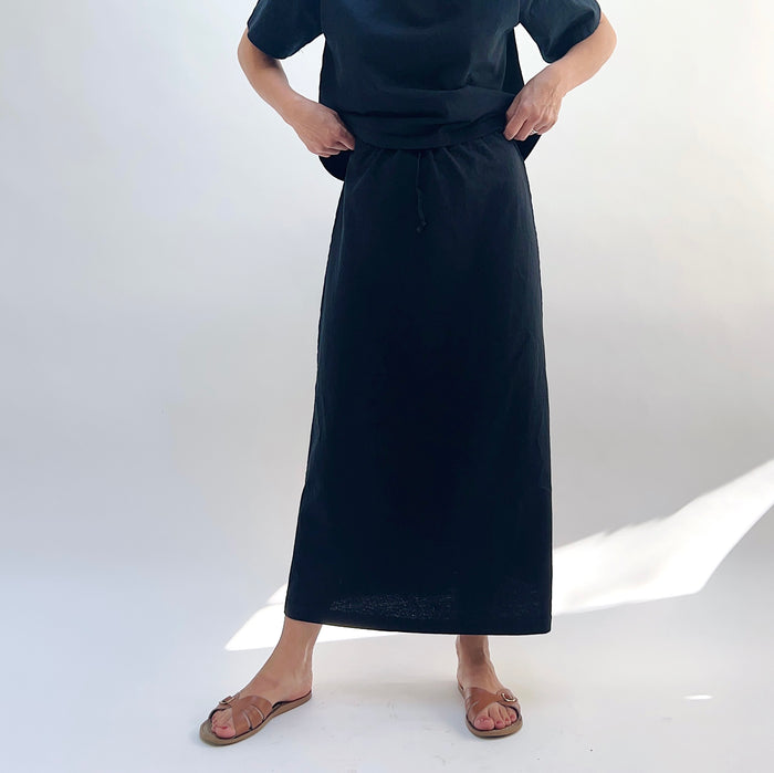 A person standing against a white background, wearing a black drawstring skirt with a waistband and a pair of sandals.