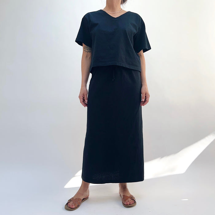 A person standing against a white background, wearing a black drawstring skirt with a waistband and a pair of sandals.