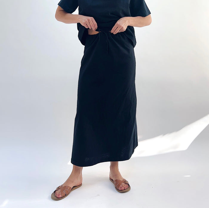 A person standing against a white background, wearing a black drawstring skirt with a waistband and a pair of sandals.