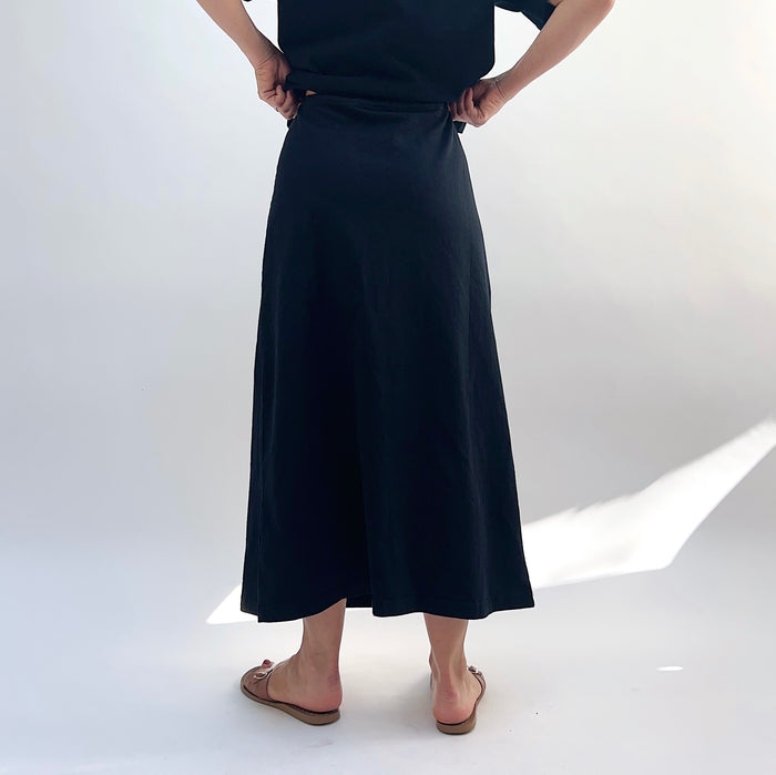 A person standing against a white background, wearing a black drawstring skirt with a waistband and a pair of sandals, viewed from the back