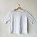 A white elbow sleeve top hanging on a wooden hanger against a textured wall.
