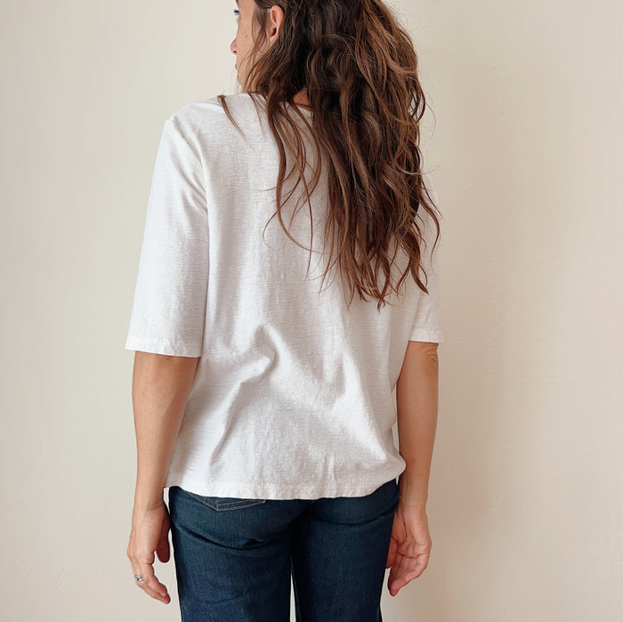 A person standing against a white background, wearing a white elbow sleeve top and blue jeans, viewed from the back