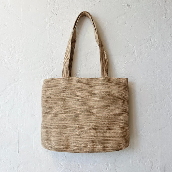 Dotter medium tan tweed tote bag with woven raffia texture and shoulder straps, hanging against a white wall