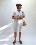 Woman wearing a white dress with buttons down the front and holding a woven bag against a white background