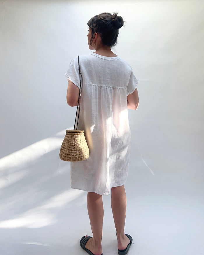 Person wearing a white dress holding a woven bag against a white background, viewed from behind