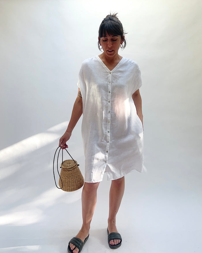 Woman wearing a white dress with buttons down the front and holding a woven bag against a white background