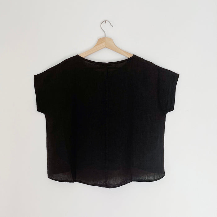 A black high-low hem linen tee shirt with a boat neckline, displayed on a wooden hanger against a white background, viewed from behind
