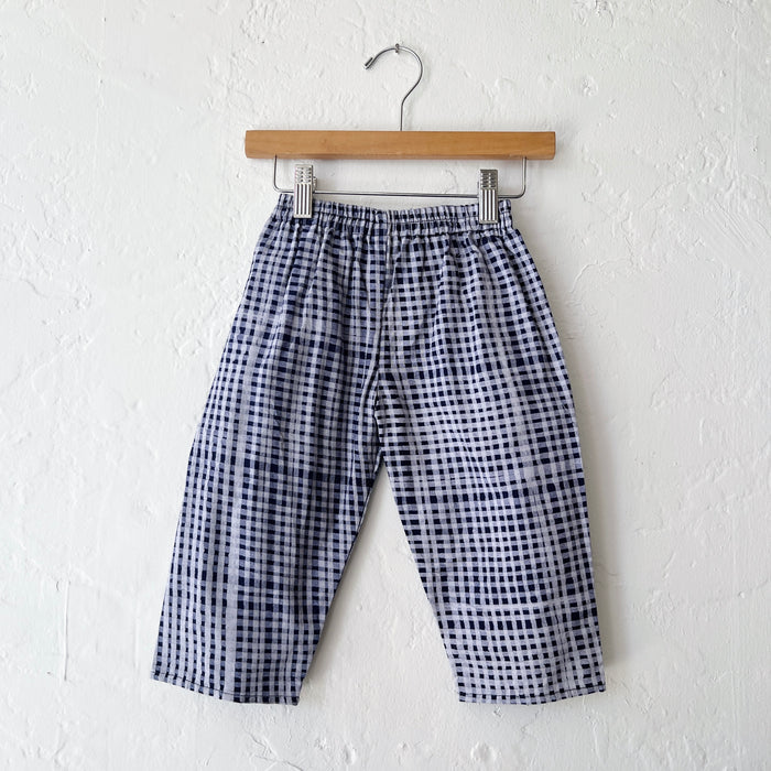 Kids lazy pants in indigo gingham Batiki cotton, hanging flat on a wooden clip hanger against a white wall, showing elasticated waistband and relaxed fit