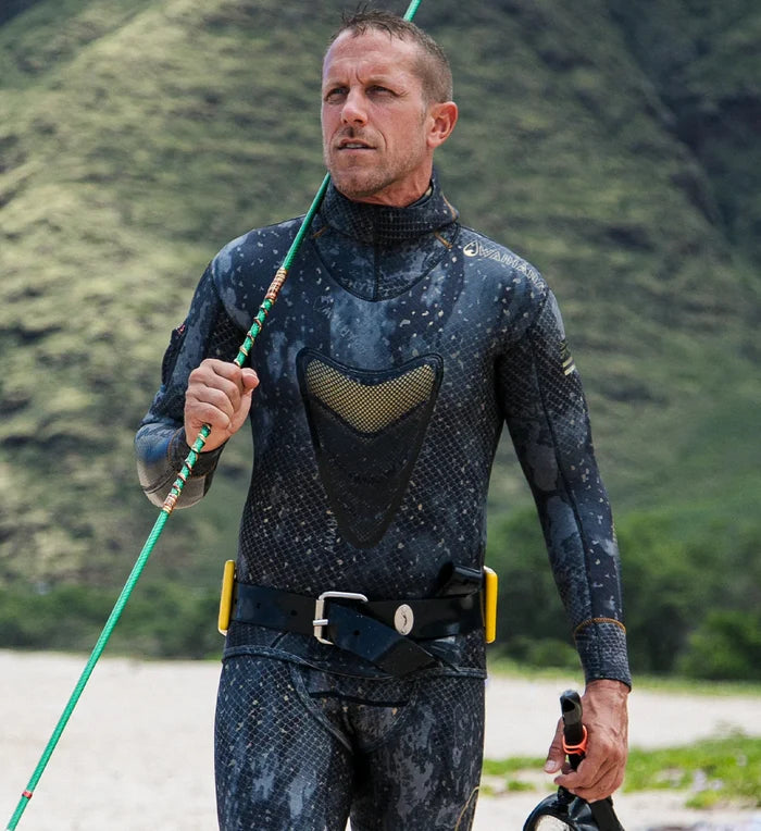 Male spearfisher wearing a Waihana camouflage wetsuit and Lava Rock weight belt, holding a pole spear and fins on a sandy beach with lush green mountains behind him.