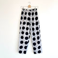 Full front view of Lazy Pants in Indigo Dots Batiki cotton, hanging on a clip hanger against a white wall, showing elastic waistband, bold hand-printed oversized black polka dots on a grey-white ground, and relaxed wide-leg silhouette
