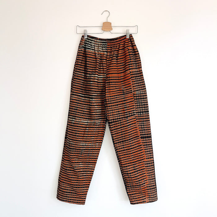 Lazy Pants in Persimmon Grid — full-length front view on hanger, loose-fit unisex pants in hand-dyed persimmon orange and black batik stripe print with elastic waist