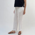 A person standing against a white background, wearing white linen pants and a black top, with tan sandals.