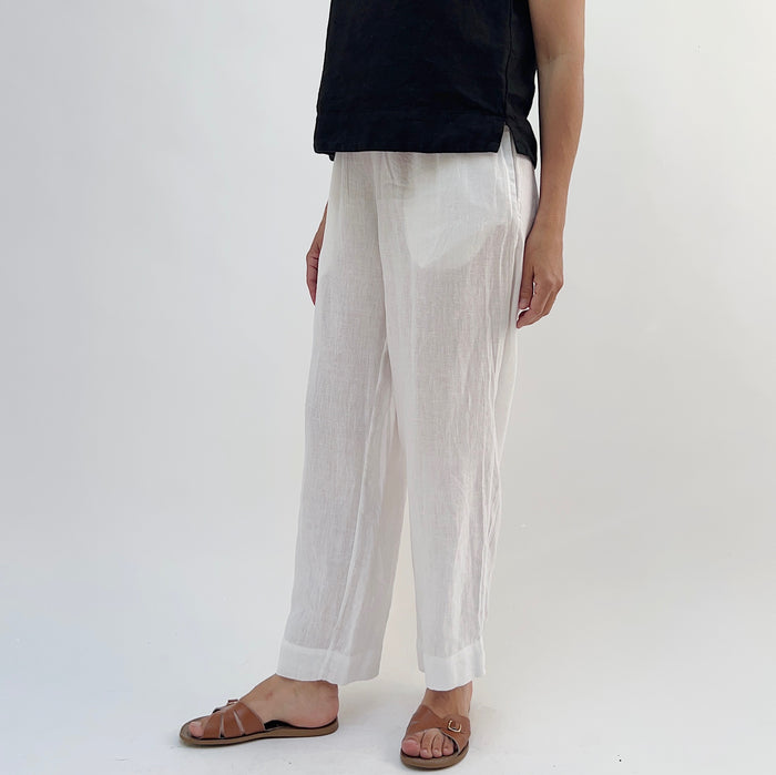 A person standing against a white background, wearing white linen pants and a black top, with tan sandals.
