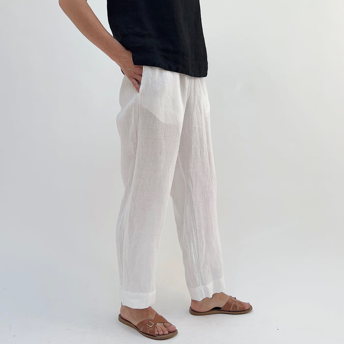 A person standing against a white background, wearing white linen pants and a black top, with tan sandals.