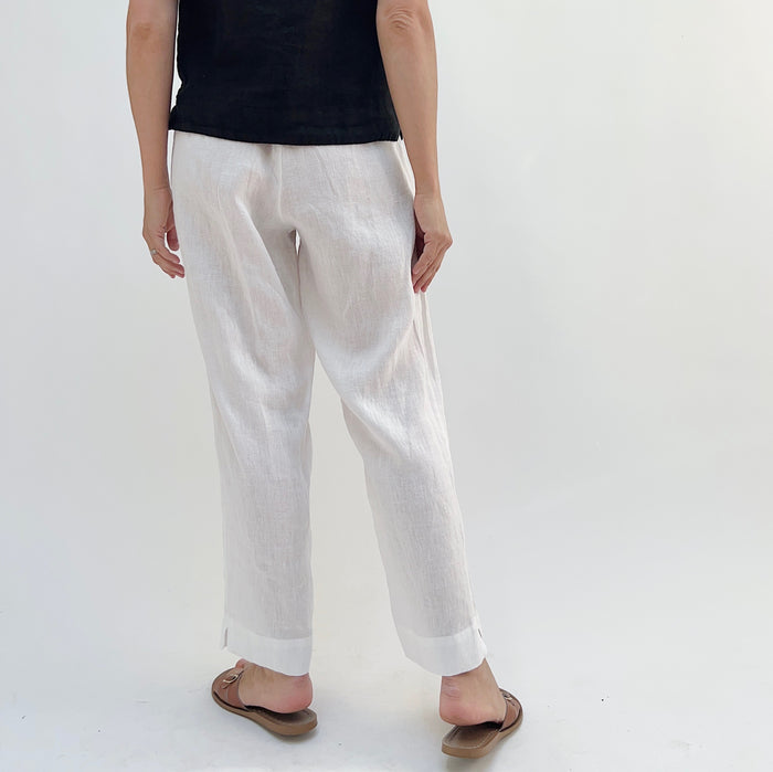 A person standing against a white background, wearing white linen pants and a black top, with tan sandals, viewed from the back