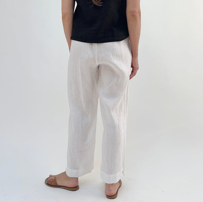 A person standing against a white background, wearing white linen pants and a black top, with tan sandals, viewed from the back