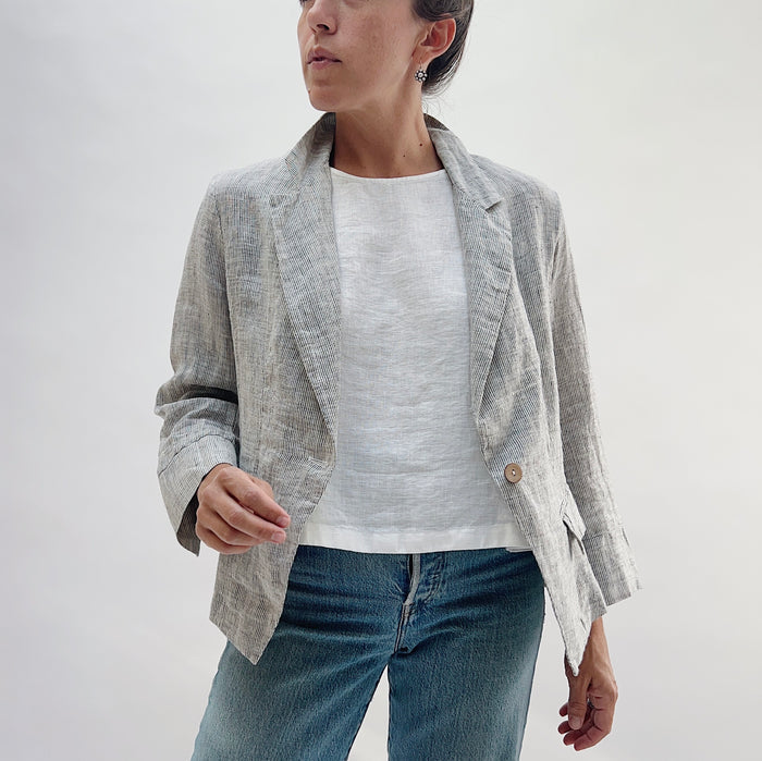 Front three-quarter view of the Linen Blazer in Grey Pinstripe by Yuvita, showing the open lapel, single-button closure, and flap pockets, styled with a white linen tee and blue jeans
