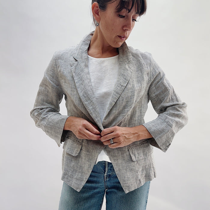 Front view of the Linen Blazer in Grey Pinstripe by Yuvita being buttoned, showing the single-button closure, notched lapel, and flap pockets, styled with a white linen tee and blue jeans