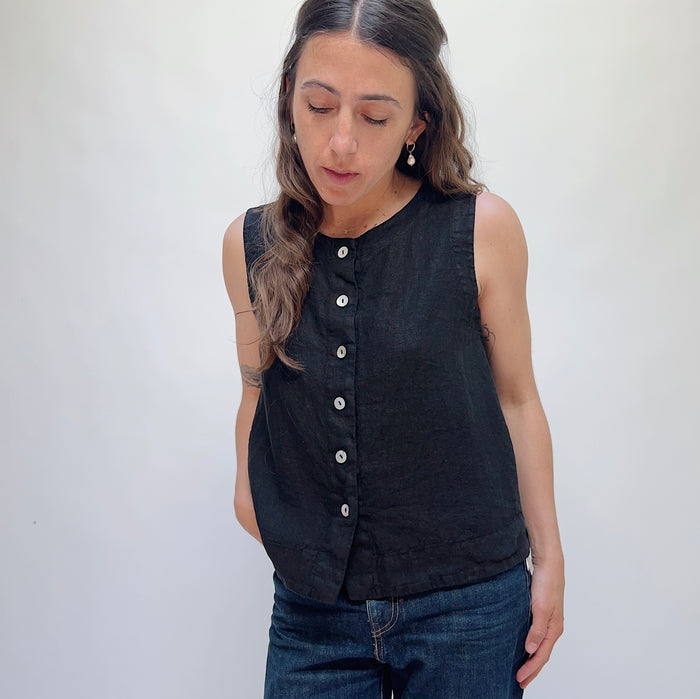 A person wearing a black linen button down tank top and blue jeans standing against a white background.