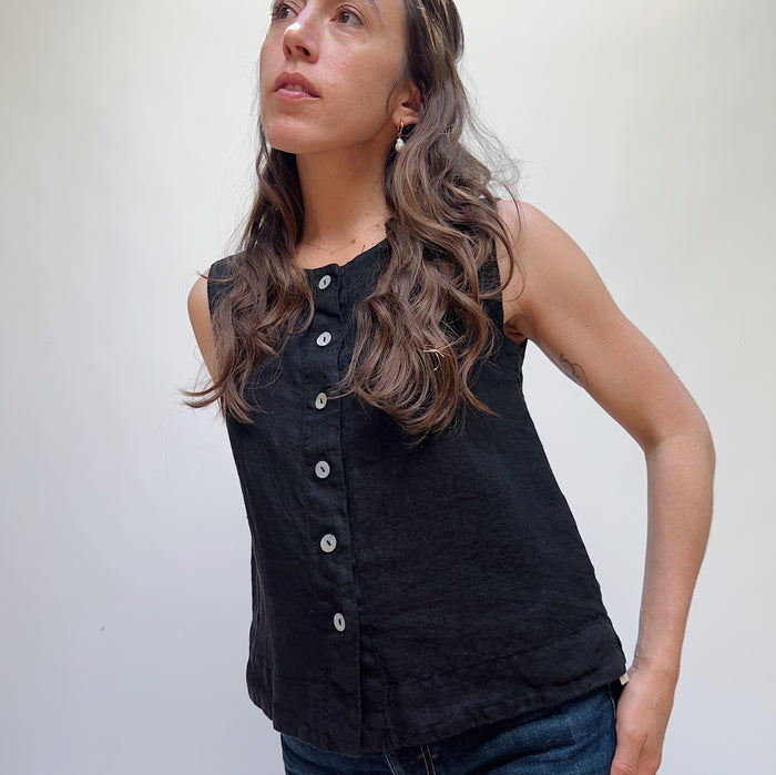 A person wearing a black linen button down tank top and blue jeans standing against a white background.