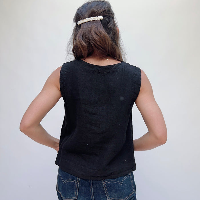 A person wearing a black linen button down tank top and blue jeans standing against a white background, viewed from the back