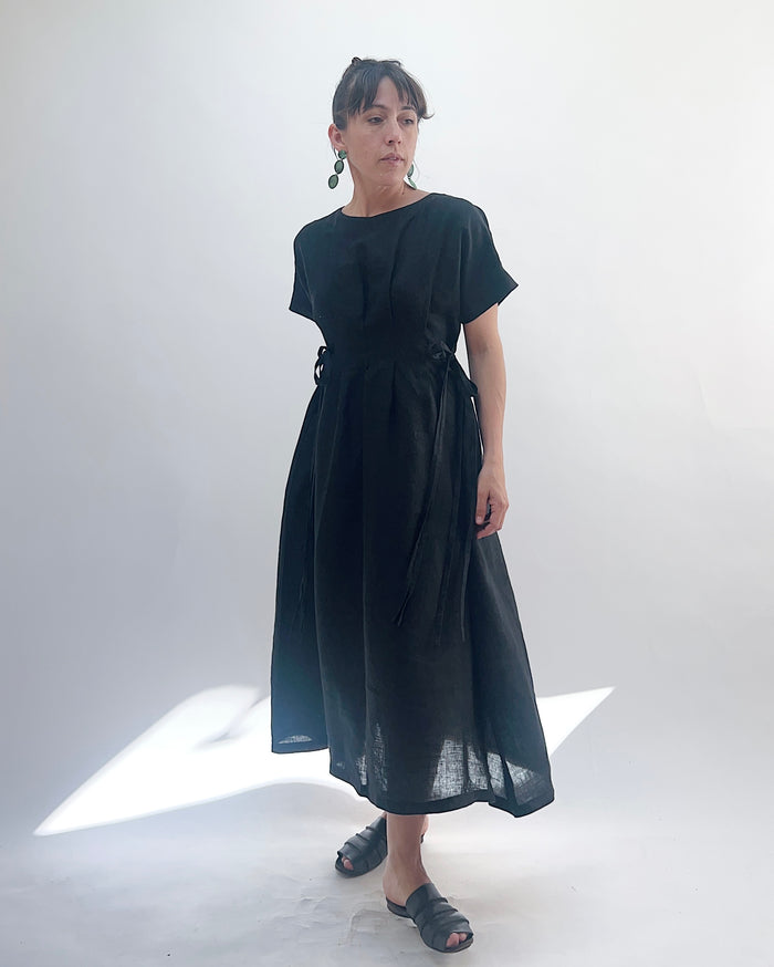 Woman with dark hair in a bun wearing a black linen dress and green dangle earrings standing against a white background.
