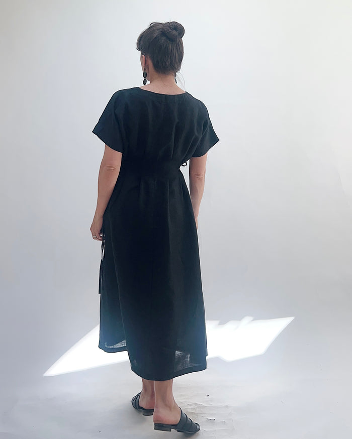 Woman with dark hair in a bun wearing a black linen dress and green dangle earrings standing against a white background as seen from behind.