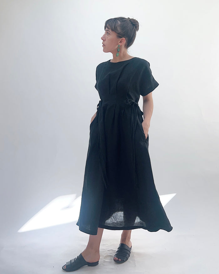 Woman with dark hair in a bun wearing a black linen dress and green dangle earrings standing against a white background.