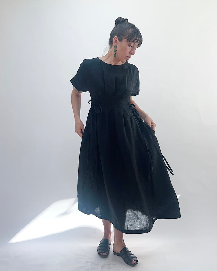 Woman with dark hair in a bun wearing a black linen dress and green dangle earrings standing against a white background.