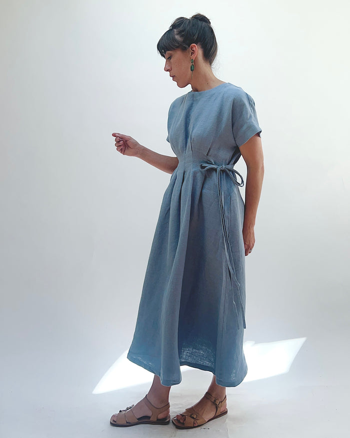 Woman with dark hair in a bun wearing a light blue linen dress and green dangle earrings standing against a white background.