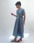 Woman with dark hair in a bun wearing a light blue linen dress and green dangle earrings standing against a white background.