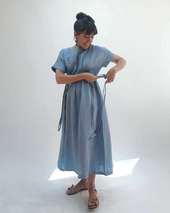 Woman with dark hair in a bun wearing a light blue linen dress and green dangle earrings standing against a white background tying the dress strings at one side of her body.