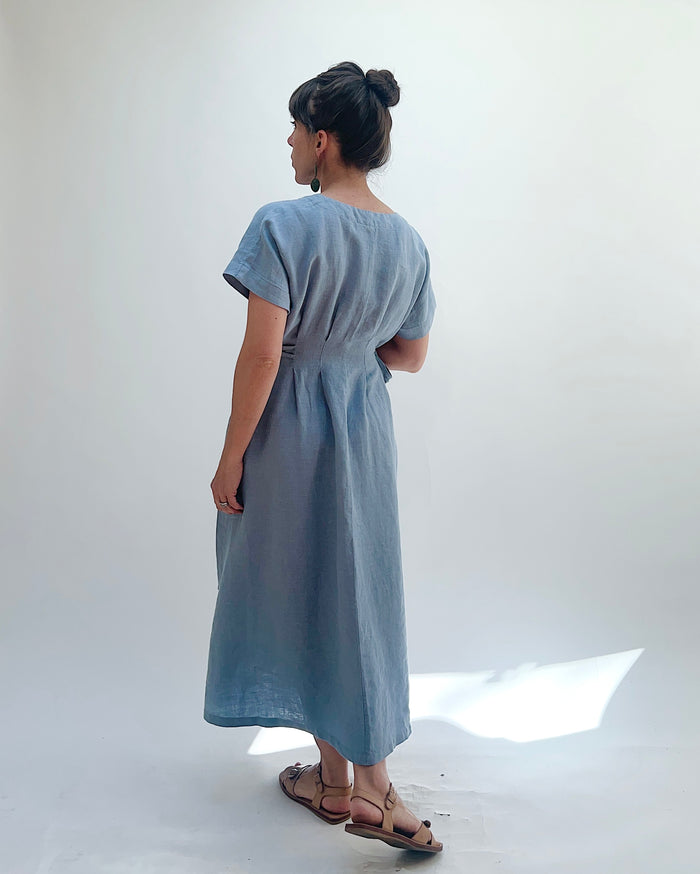 Woman with dark hair in a bun wearing a light blue linen dress and green dangle earrings standing against a white background as seen from behind.
