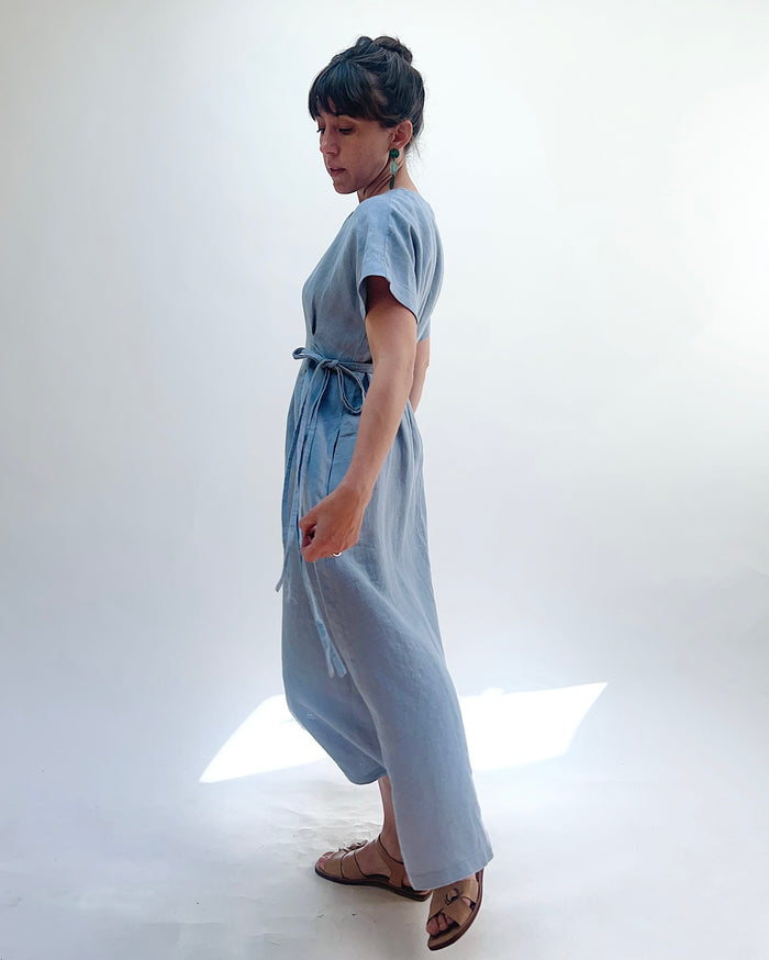 Woman with dark hair in a bun wearing a light blue linen dress and green dangle earrings standing against a white background as seen turning to the side.