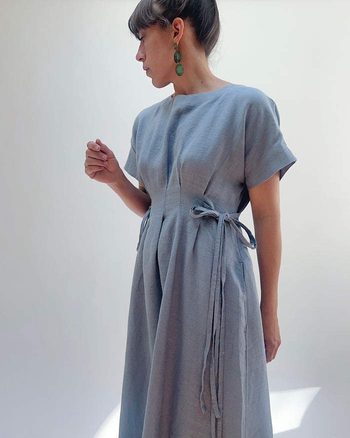 Close up of the torso of a woman with dark hair in a bun wearing a light blue linen dress and green dangle earrings standing against a white background.