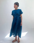 Woman with dark hair in a bun wearing a blue linen dress and green dangle earrings standing against a white background.