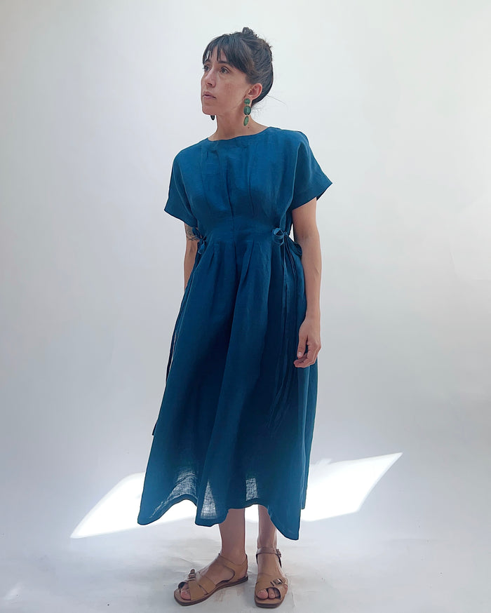 Woman with dark hair in a bun wearing a blue linen dress and green dangle earrings standing against a white background.