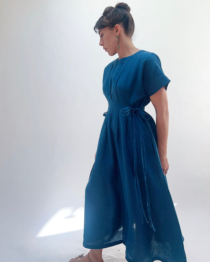 Woman with dark hair in a bun wearing a blue linen dress and green dangle earrings standing against a white background.