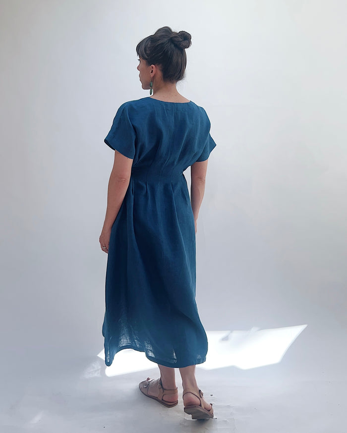 Woman with dark hair in a bun wearing a blue linen dress and green dangle earrings standing against a white background as seen from behind.