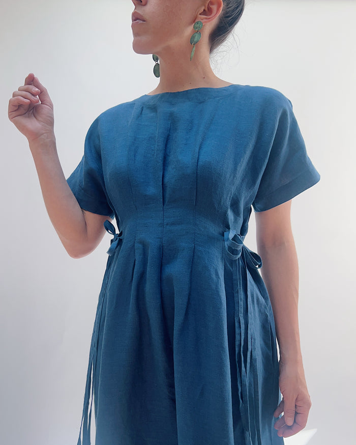 Close up view of a woman with dark hair in a bun wearing a blue linen dress and green dangle earrings standing against a white background.
